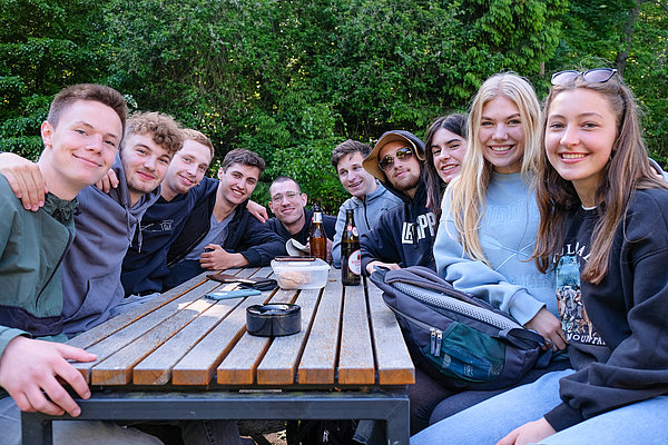 Eine Gruppe von zehn jungen Menschen sitzt an einem Holztisch im Freien. Sie lächeln und stehen eng zusammen, einige haben   Getränke vor sich. Die Umgebung ist grün mit Bäumen im Hintergrund.