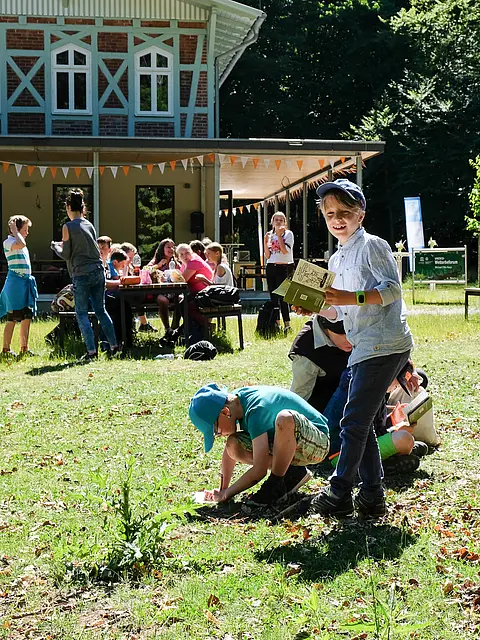 Kinder spielen im Freien, einige graben im Boden, während andere am Tisch sitzen und essen. Ein Junge lächelt und hält ein Buch in der Hand, umgeben von Bäumen und einer rustikalen Gebäude im Hintergrund. Die Atmosphäre ist fröhlich und lebhaft.