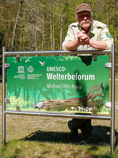 Ein Mann in Naturbekleidung steht hinter einem Schild, das "UNESCO-Welterbe­forum" und "Michael Otto Haus" anzeigt. Das Schild ist grün gestaltet und zeigt einige Illustrationen von Pflanzen und Tieren. Im Hintergrund ist eine waldreiche Umgebung sichtbar.