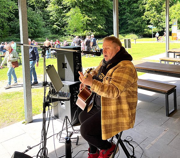 Ein Musiker sitzt auf einem Stuhl in einem Pavillon und spielt Gitarre. Er trägt eine gemusterte Jacke und hat einen Mikrofonständer neben sich. Im Hintergrund sind Zuschauer auf einer Wiese zu sehen, während Bäume den Raum umgeben.