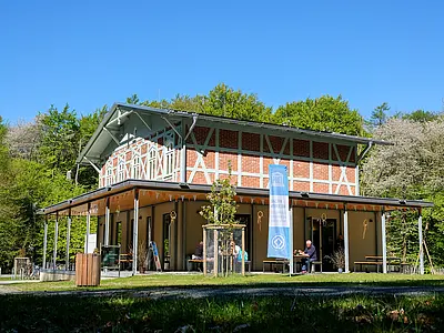 Ein modernes Gebäude im Freien, umgeben von Bäumen. Vor dem Gebäude befinden sich einige Tische und Bänke, an denen Menschen sitzen. Eine blaue Fahne mit einem Logo ist sichtbar. Der Himmel ist klar und sonnig, was eine einladende Atmosphäre schafft.